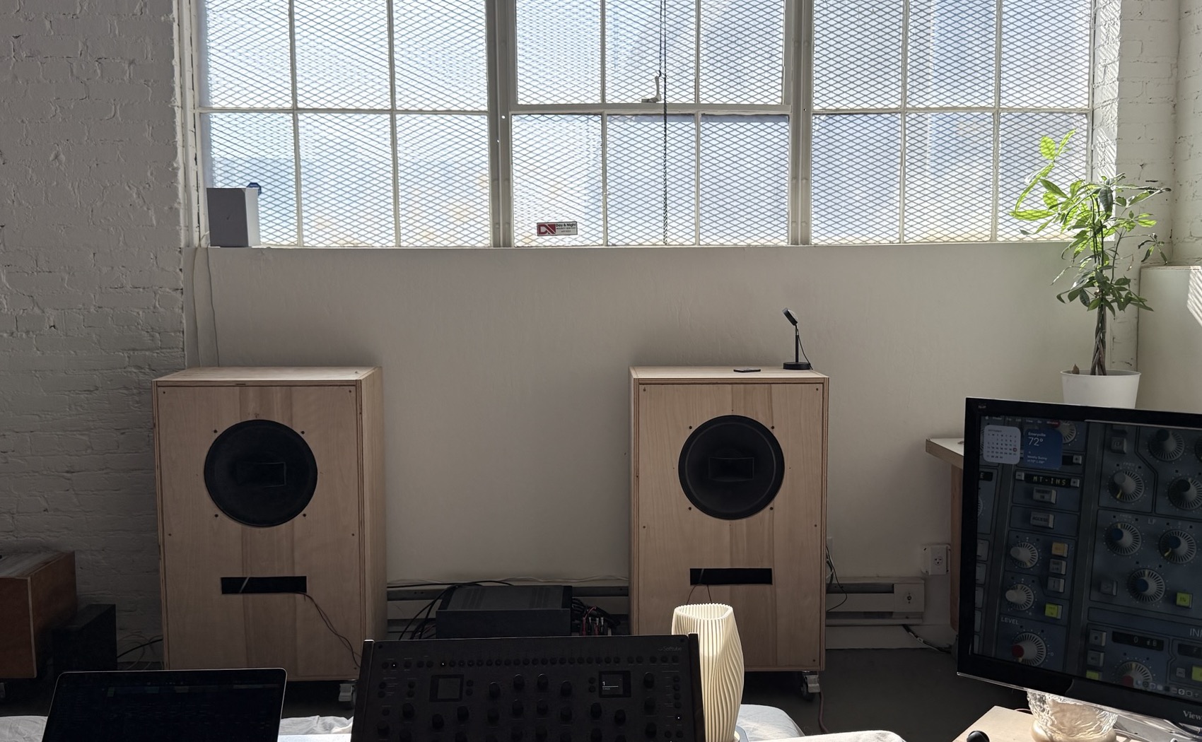 Two tall light-plywood speaker cabinets, each with one large round driver and a slot port, standing under the studio's steel-framed window.