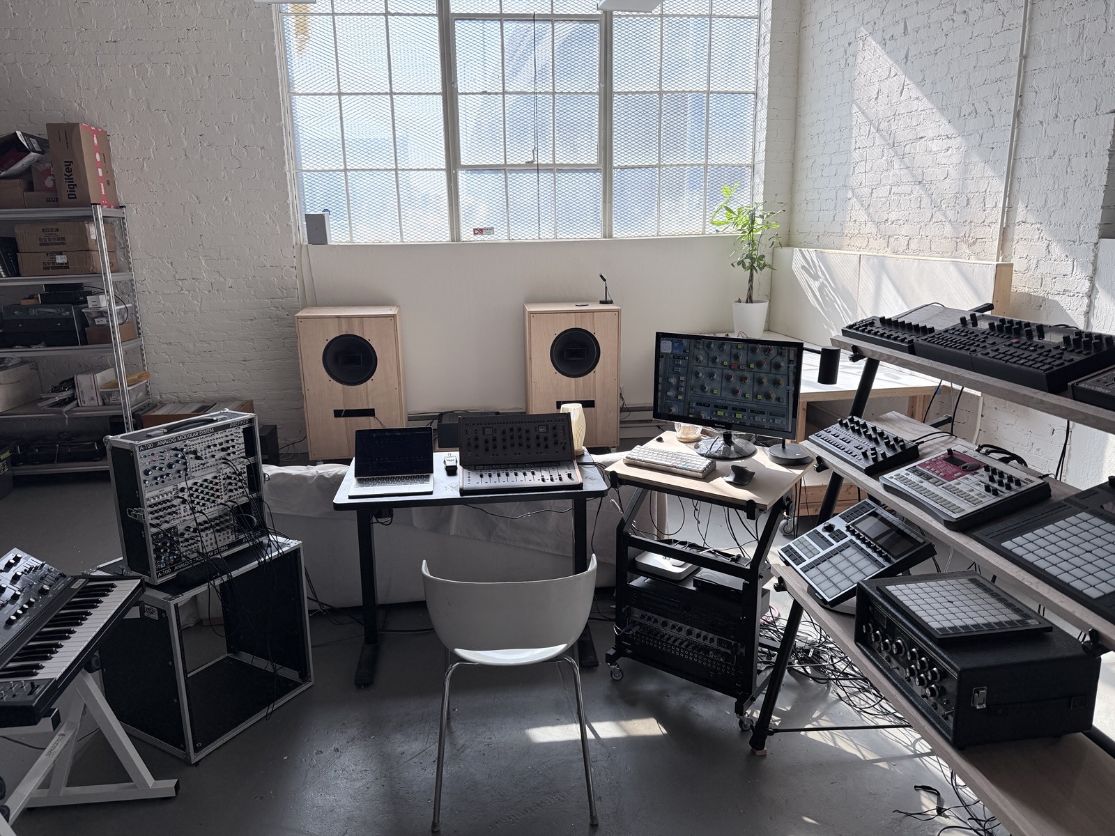 The studio: a modular case and a synth on a stand at left, a desk with a laptop and a channel-strip controller, a pair of plywood speakers under a tall industrial window, and a tiered stand of drum machines and controllers at right.